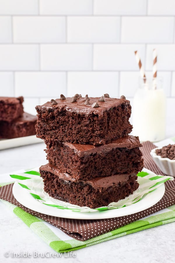 A white plate with three frosted zucchini brownies stacked on it and glass of milk behind it.