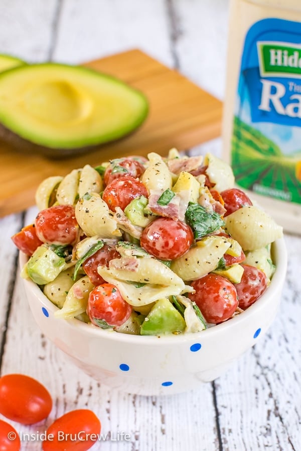 A white bowl on a white background filled with ranch BLT pasta salad and cutting board with avocados behind it