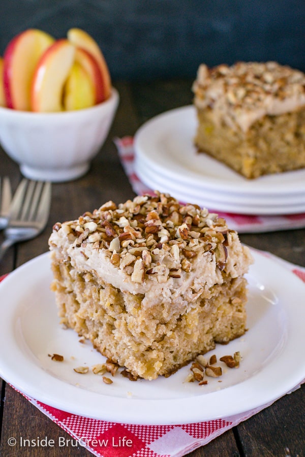 A white plate with a piece of apple praline cake topped with frosting and pecans on it