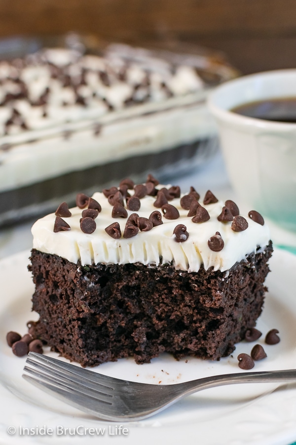 A slice of chocolate zucchini cake with frosting and chocolate chips on a white plate with a bite taken out of it.