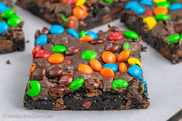 A tray with parchment paper and squares of fudge brownies with M&M's on it.