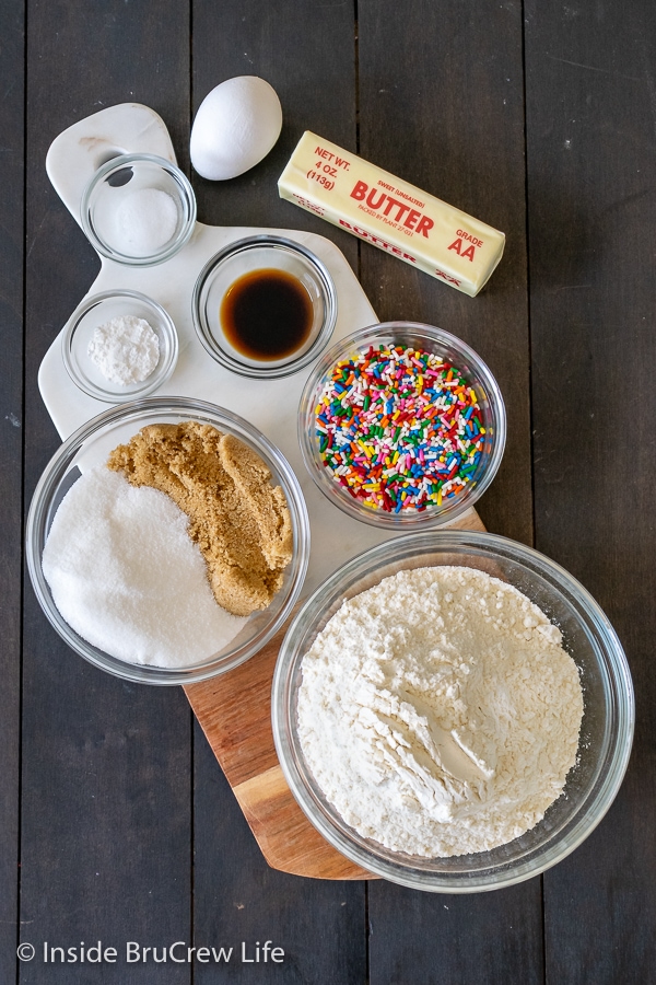 Bowls of ingredients needed to make Funfetti Sugar Cookie Bars on a brown board