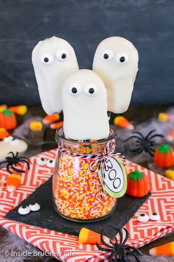 Three ghost cookies attached to straws placed in a jar of sprinkles.
