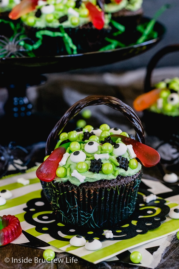 Brownie cupcake topped with green frosting and spooky sprinkles on a black doily with more cupcakes behind it