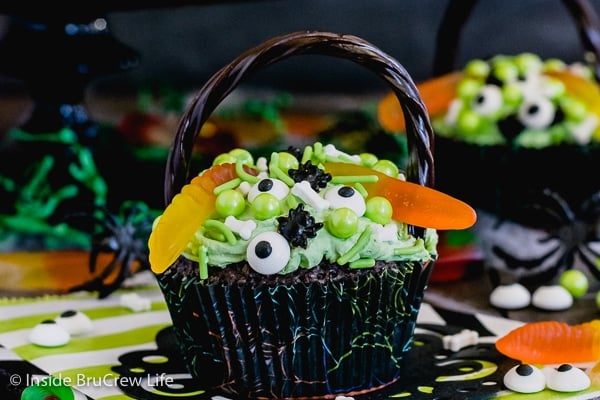 A close up picture of a brownie cupcakes topped with green frosting, gummy worms, and spooky sprinkles to look like a witch's cauldron