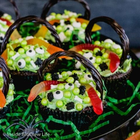 A black cake stand with brownie cupcakes decorated with candies and gummy worms to look like witch cauldrons