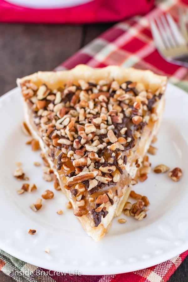A white plate with a slice of apple cheesecake tart topped with caramel, chocolate, and pecans on it.