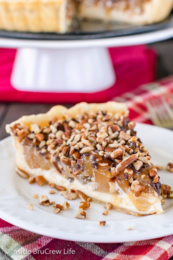 A white plate with a slice of apple cheesecake tart topped with pecans on it.