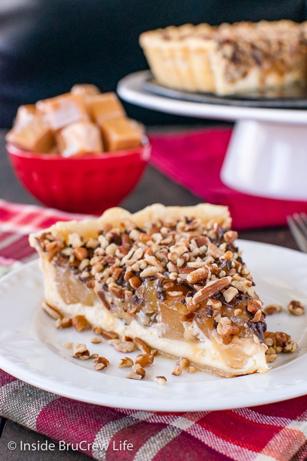 A white plate with a slice of caramel apple tart topped with pecans on it.