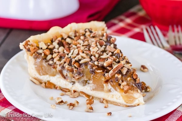 A white plate with a slice of apple cheesecake pie topped with caramel, chocolate, and nuts.