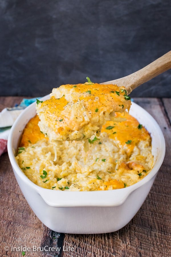 A white casserole dish filled with cheesy hashbrown casserole with a spoon lifting some of the potatoes out.