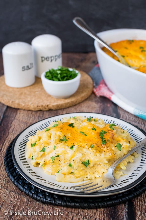 A white plate with a serving of cheesy hashbrowns on it and a casserole dish behind it.