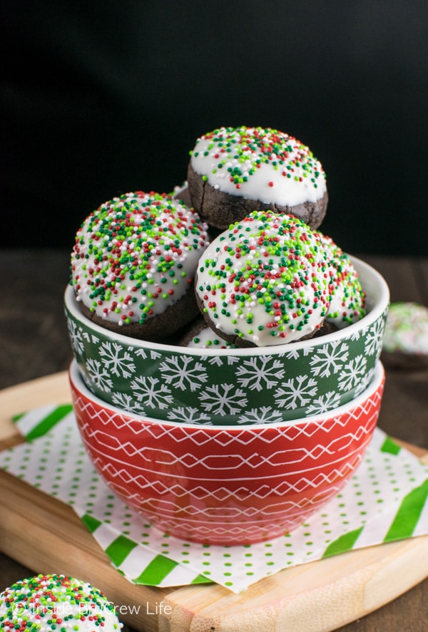 A red and green bowl on a wood tray filled with chocolate mint snowball cookies topped with chocolate and sprinkles