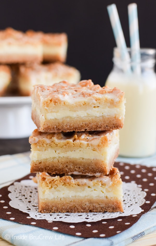 A white doily with three eggnog cheesecake crumble bars stacked on it and a bottle of eggnog behind it