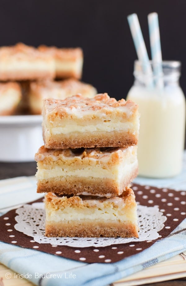 A white doily with three eggnog cheesecake bars stacked on it and a plate of cheesecake bars and eggnog behind it