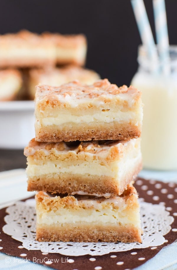 Three eggnog cheesecake bars stacked on a white doily with a plate of more cheesecake bars and eggnog behind it