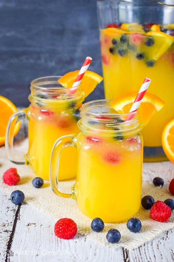 Two mugs filled with pineapple punch and floating berries with a pitcher of punch behind them