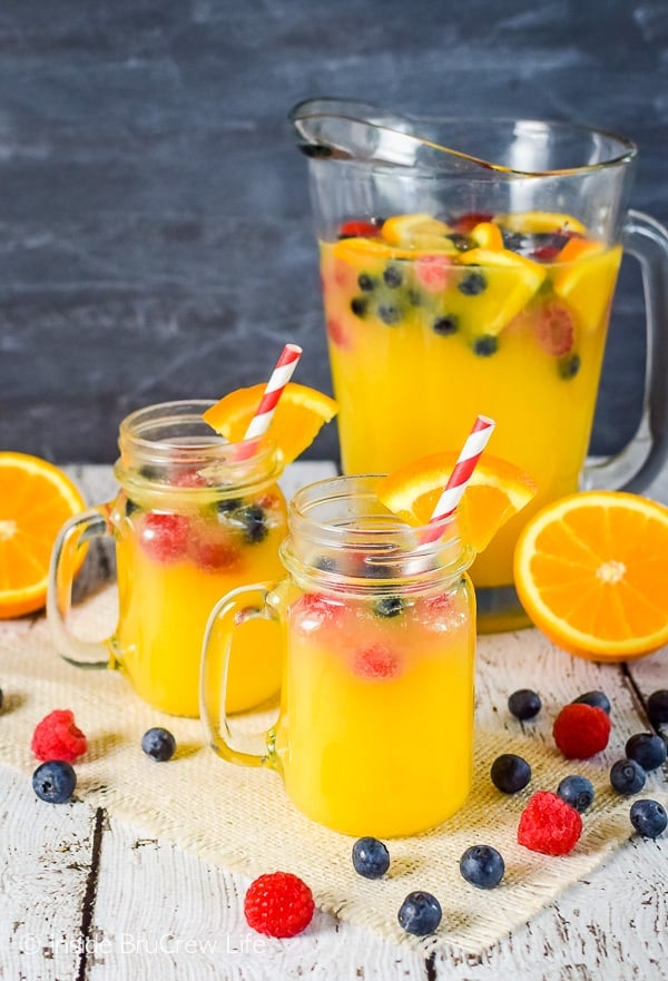 Two clear mugs with pineapple orange juice punch and fresh berries with a pitcher of punch behind them