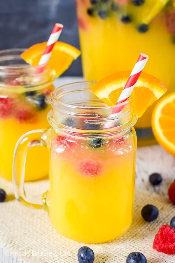 A clear mug with pineapple punch and floating fresh berries in it and a pitcher of juice behind it