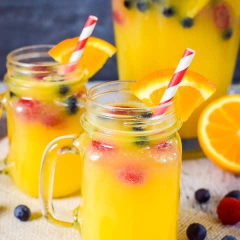 Two clear glasses filled with pineapple punch and fresh berries on a tan towel and more berries around the mugs