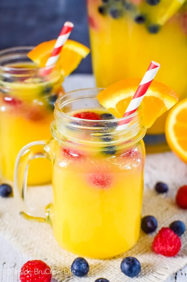 A clear mug filled with pineapple punch and floating berries and served with red striped straw and orange slice