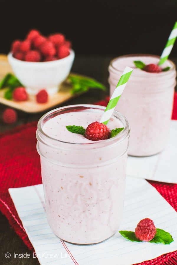 A pink smoothie in a clear mason jar with a raspberry, mint leaf, and green and white straw in it.