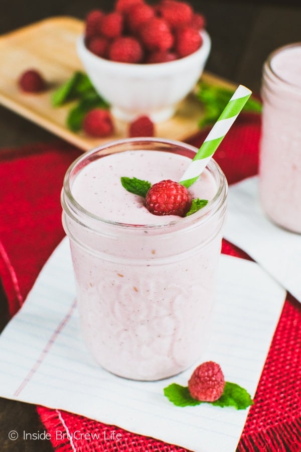 A pink smoothie in a clear mason jar with a raspberry, mint leaf, and green and white straw in it.