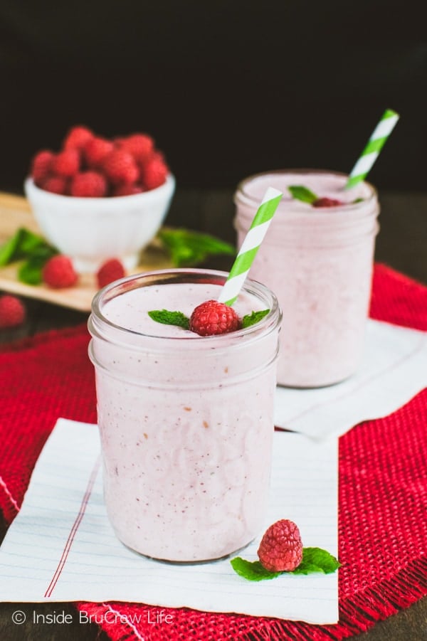 A pink smoothie in a clear mason jar with a raspberry, mint leaf, and green and white straw in it.