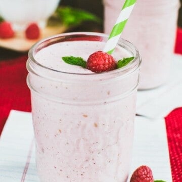 A pink smoothie in a clear mason jar with a raspberry, mint leaf, and green and white straw in it.