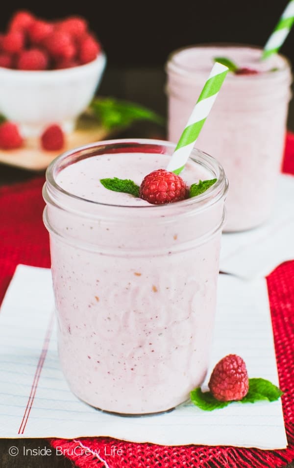 A pink smoothie in a clear mason jar with a raspberry, mint leaf, and green and white straw in it.