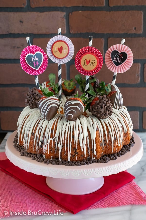 A full sized Strawberry Chocolate Chip Bundt Cake on a pink cake plate with paper straws and flag decorations on top