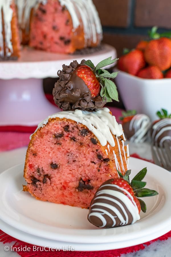 A slice of strawberry bundt cake on a white plate with a chocolate covered strawberry on top and beside it