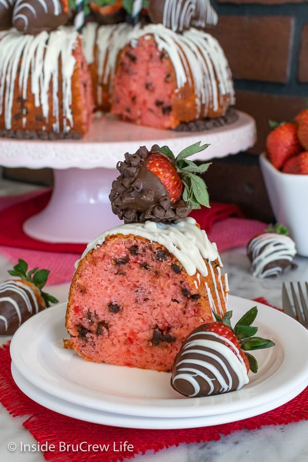 A white plate with a slice of strawberry chocolate chip bundt cake with chocolate covered strawberries and the cake behind it