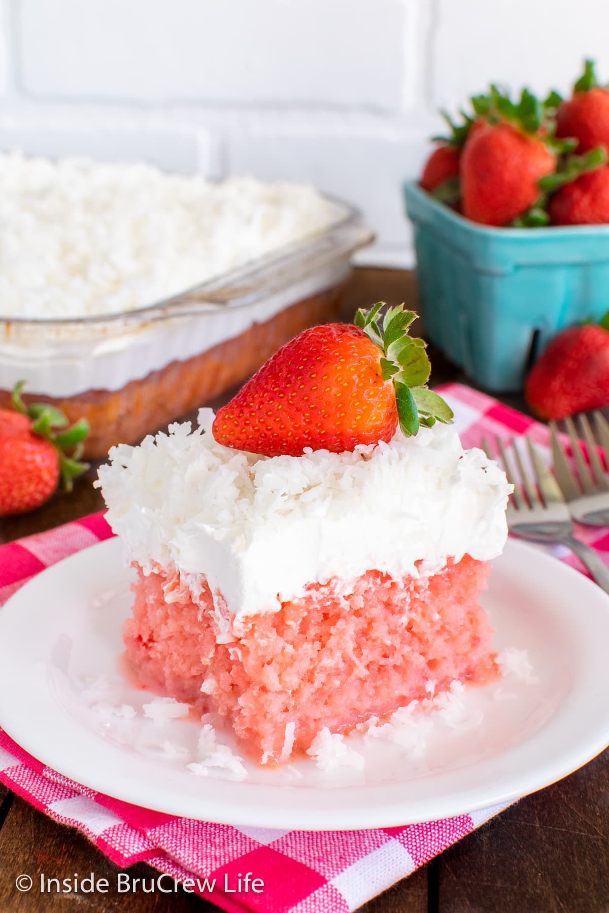 A white plate with a slice of pink strawberry cake topped with Cool Whip and coconut.