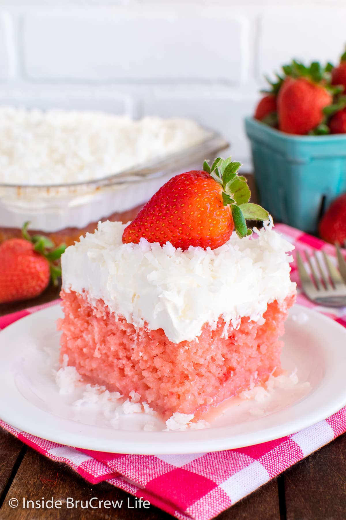 A slice of strawberry poke cake on a white plate topped with cool whip and coconut.