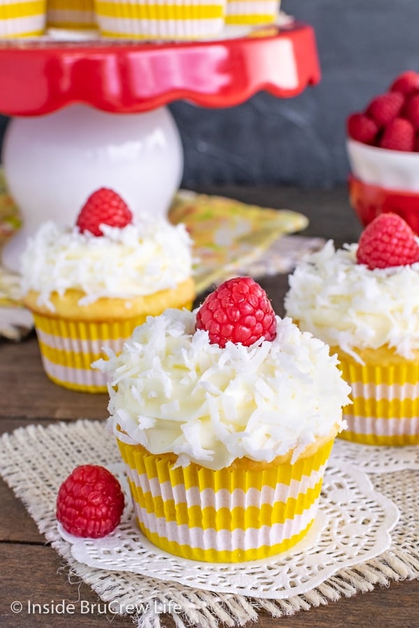 Three coconut cupcakes with yellow and white liners topped with lemon frosting, shredded coconut, and a raspberry on a wooden board.