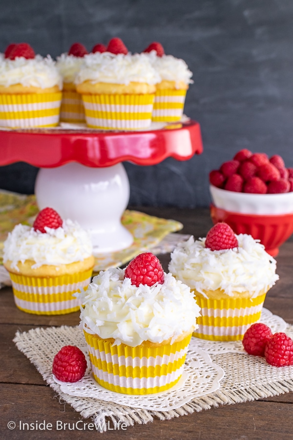 Three lemon coconut cupcakes topped with shredded coconut and raspberries on a wooden board and a cake plate with more cupcakes behind them.