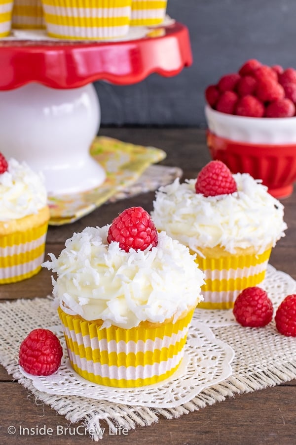 Lemon coconut cupcakes with lemon frosting, shredded coconut, and fresh raspberries on a wooden board with a bowl of more berries.