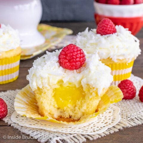 Two coconut cupcakes topped with lemon frosting and coconut and a bite out of the front one showing a lemon filling center.