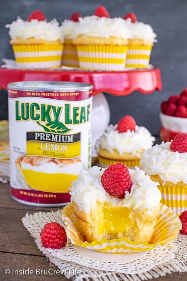 A can of lemon pie filling with lemon coconut cupcakes all around it. One cupcake has a bite taken out showing the hidden lemon center.