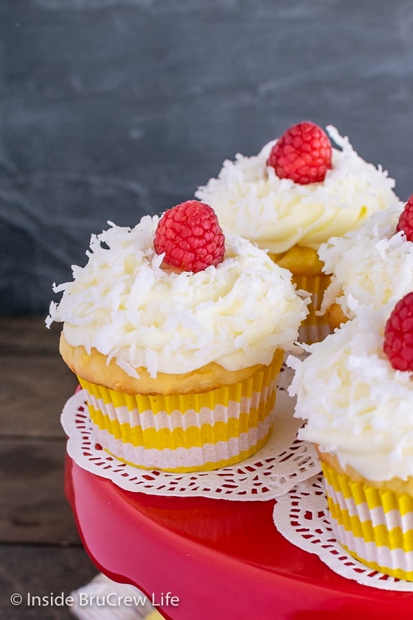 A red cake plate with coconut cupcakes in yellow liners topped with lemon frosting, shredded coconut, and fresh raspberries.