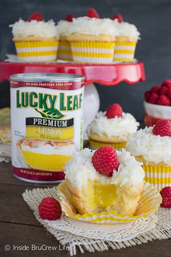 A picture of Lemon Coconut Cupcakes with a bite taken out to see the hidden lemon filling and more cupcakes on a on a cake stand