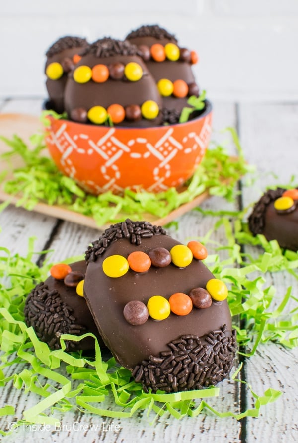 Two Reese's eggs decorated with Reese's Pieces and chocolate sprinkles on a white board with green Easter grass around them.