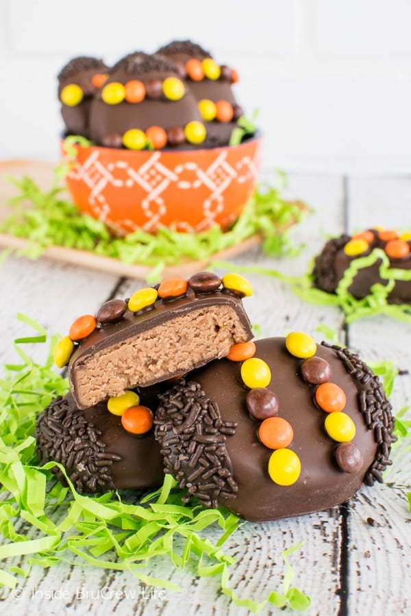 Three Homemade Reese's Eggs decorated with candy and sprinkles on a white board with green Easter grass. One egg is cut in half showing the filling.