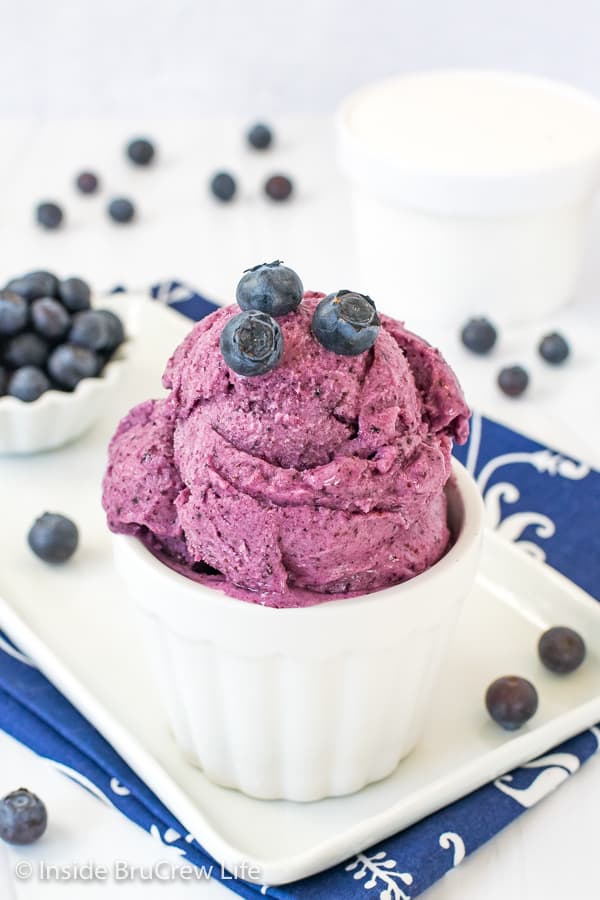 A white bowl on a white plate filled with blueberry banana frozen yogurt and topped with a few fresh blueberries