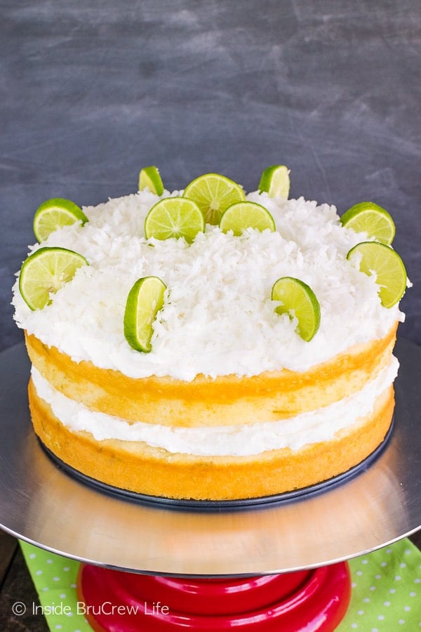 Two layers of key lime cake frosted with coconut frosting and topped with shredded coconut and key lime slices on a cake stand