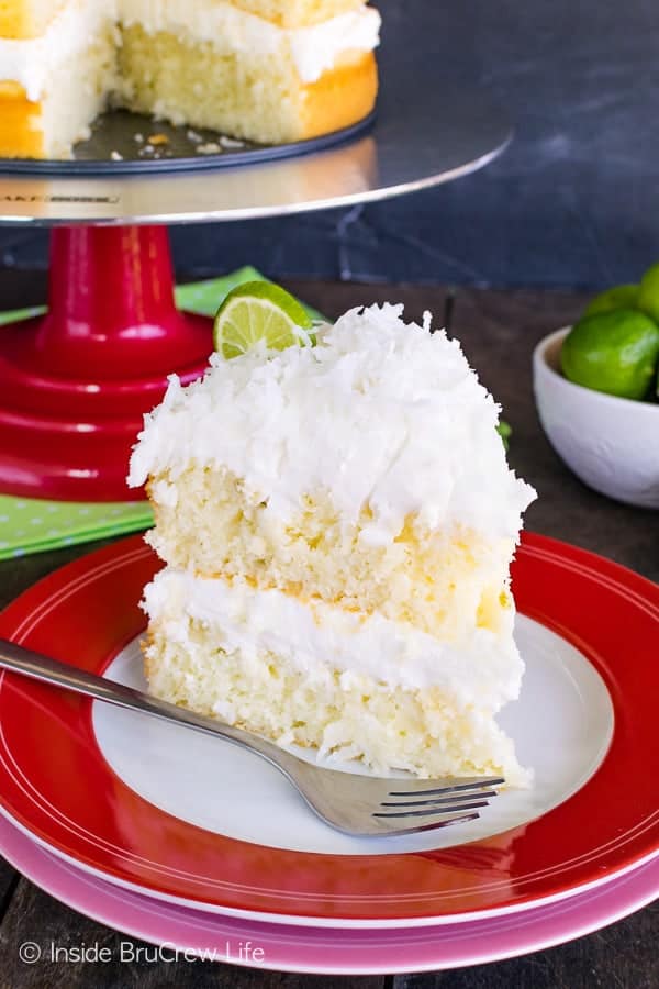 A red and white plate with a slice of key lime cake frosted with coconut frosting on it and a cake stand behind it