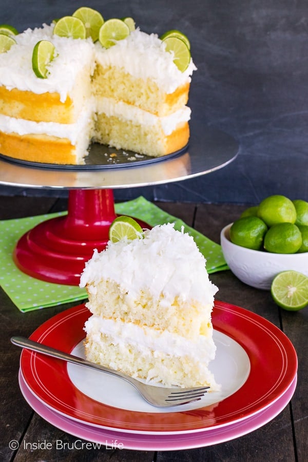 A red and white plate with a slice of coconut key lime cake with coconut frosting on it and the rest of the cake behind it on a cake stand.