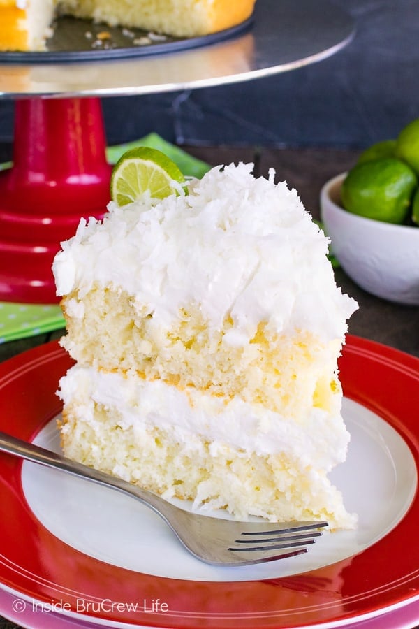 A slice of key lime cake layered with coconut frosting and topped with shredded coconut and a key lime slice on a red and white plate