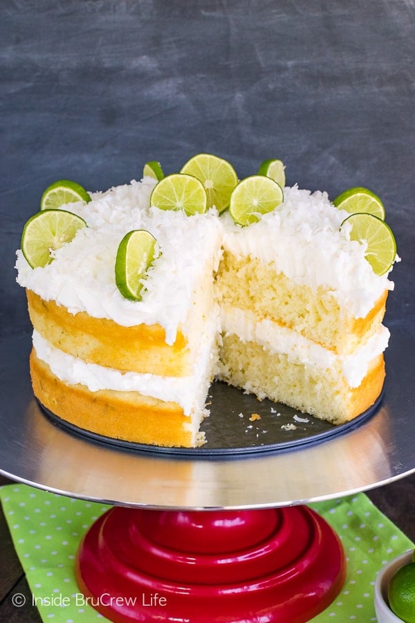 A cake stand with a key lime coconut cake decorated with shredded coconut and key lime slices on it with one slice missing.